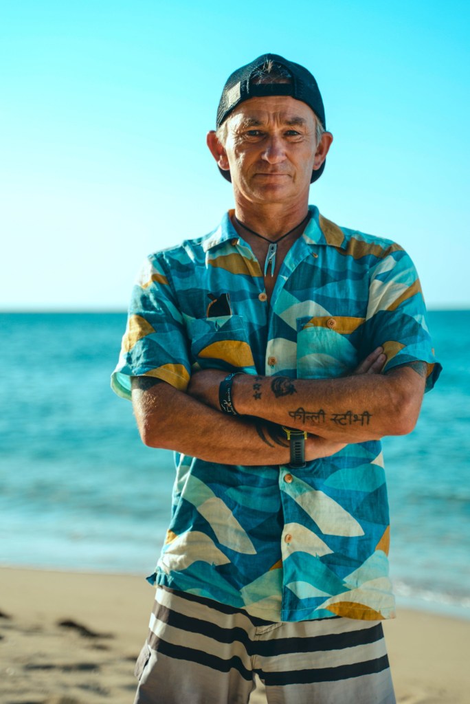 One of the contestants for the BBC reality show, Survivor called Pegleg, stood on a beach wearing a blue shirt and a backwards cap.